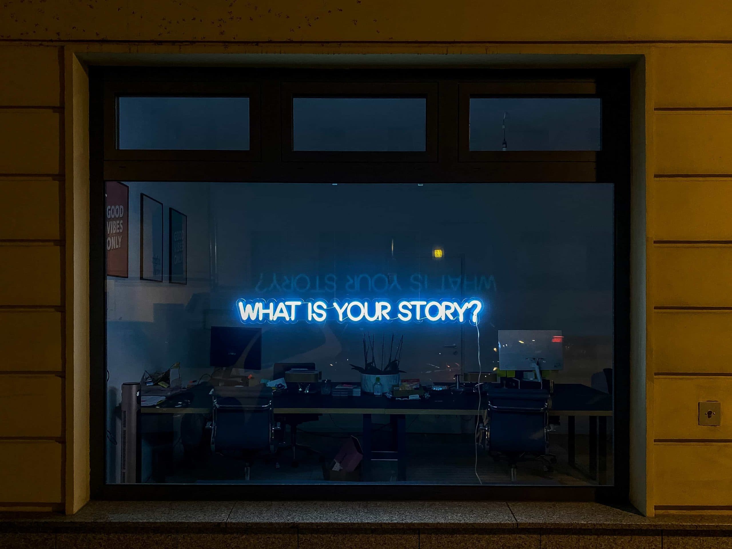 Blick von außen durch ein Fenster in ein Büro mit Leuchtschrift "What is your Story".